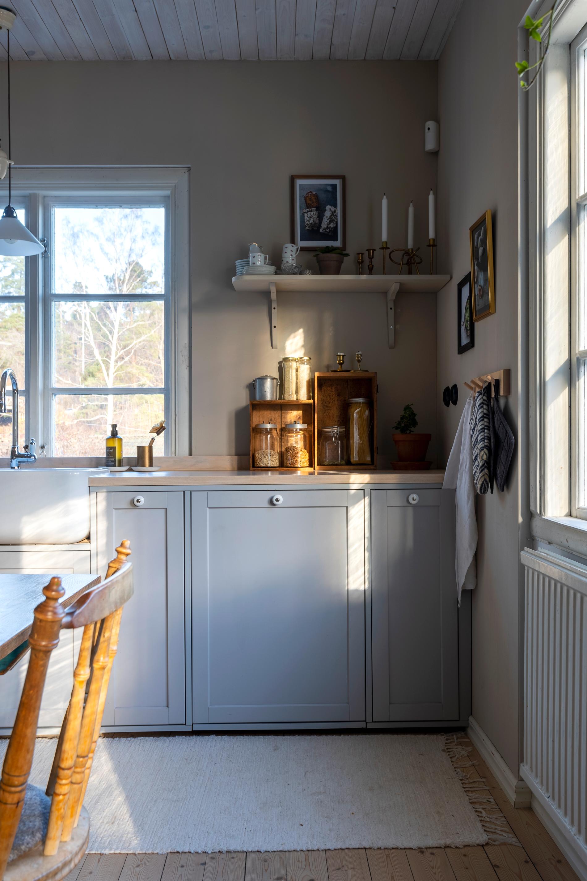 Högra hörnet av köket där det finns både ett fönster på väggen över köksskåpen och diskbänk men också ett på långsidan. De grå skåpen har en enkel profil med vita porslinsknoppar i äldre stil. På köksbänken står glasburkar med lock med olika specierivaror. 