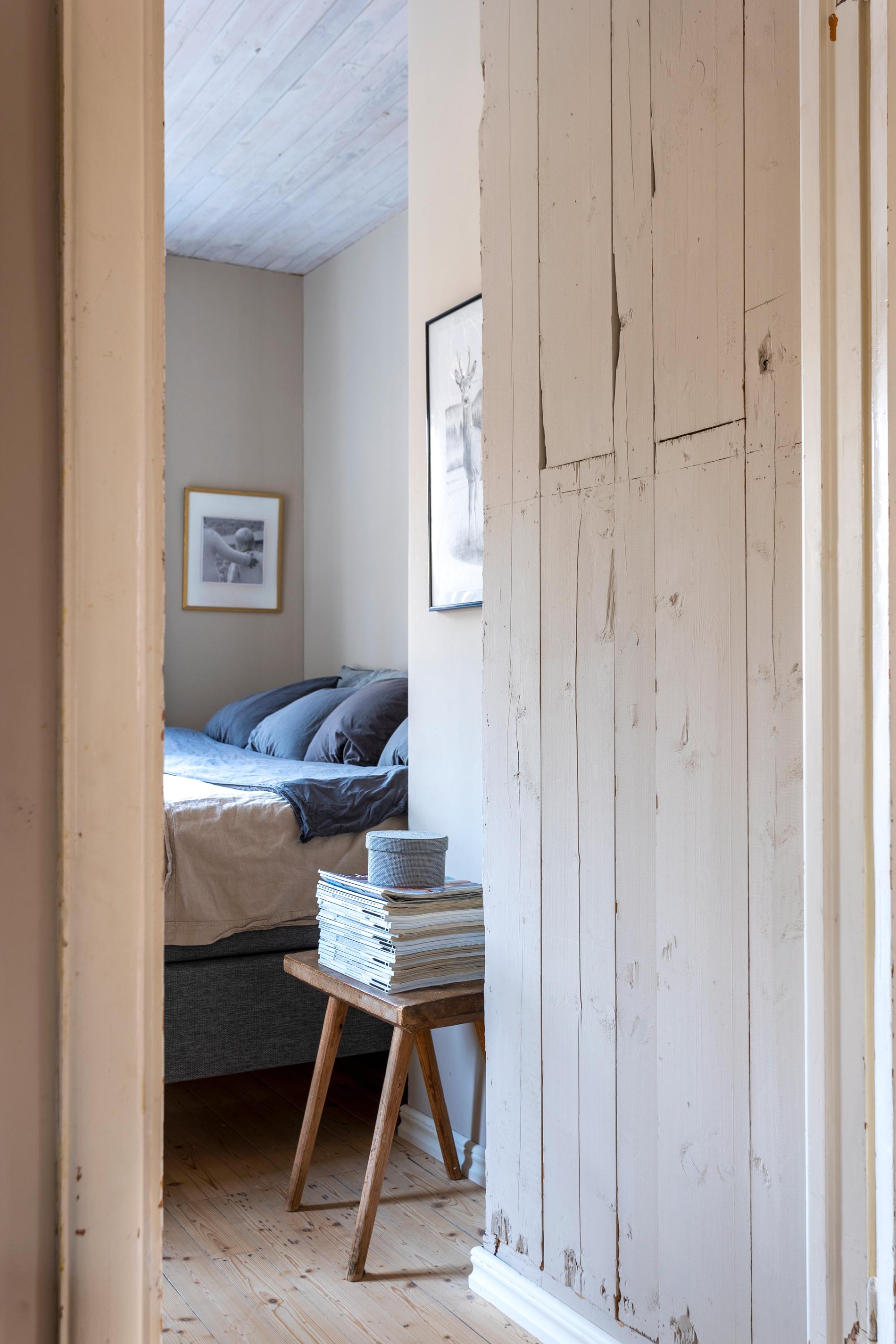 Intill föräldrasovrummet är en väggen klädd i grova plankor som målats i en ljusbeige färg. Sängen är bäddad med gråa örngott och bredvid står en rustik pall med en stor hög magasin. 