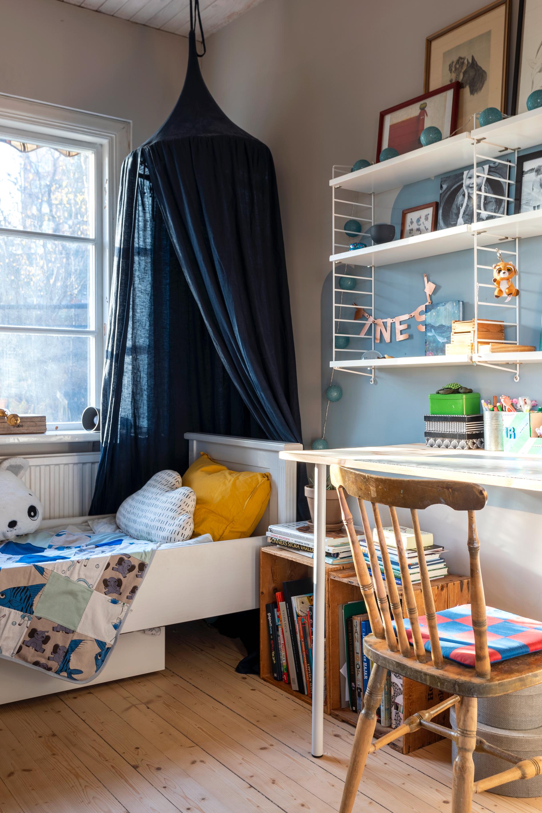 En barnsäng som står under fönstret med en mörkblå sänghimmel över som hänger över huvudänden. Över skrivbordet hänger en grundare Stringhylla i vitt. Två backar invid sängen rymmer böcker och vid skrivbordet står en lackad pinnstol av äldre modell.